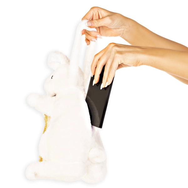 A white plush bunny handbag with gold accents, shown with a person's hands holding a phone inside its side pocket.